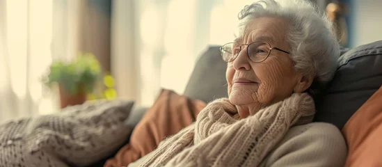 Foto op Canvas The elderly woman is sitting comfortably on a wooden couch in a cozy room, wearing glasses, a scarf, and a happy smile on her face. © TheWaterMeloonProjec