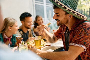 Happy man eating nacho chips while drinking beer with friends in Mexican restaurant.