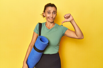 Middle aged sportswoman with yoga mat on yellow studio showing a dislike gesture, thumbs down....