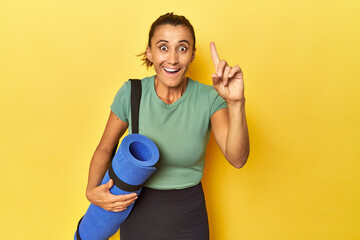 Middle aged sportswoman with yoga mat on yellow studio having an idea, inspiration concept.