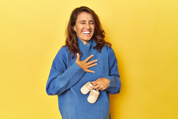 Pregnant woman holding baby shoes on yellow laughs out loudly keeping hand on chest.
