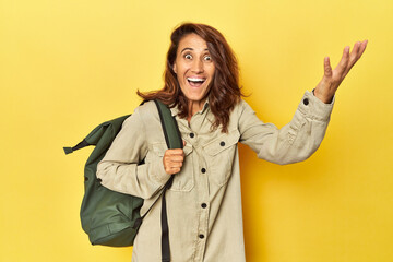 Middle aged woman ready for travel with backpack on yellow receiving a pleasant surprise, excited...