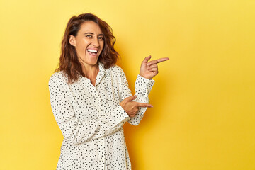 Middle-aged woman on a yellow backdrop pointing with forefingers to a copy space, expressing excitement and desire. - obrazy, fototapety, plakaty