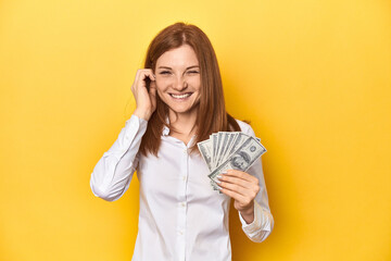 Redhead with dollar bills, cash in hand covering ears with hands.