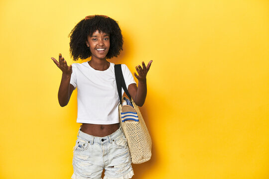 Young girl with beach bag ready for holiday, yellow backdrop receiving a pleasant surprise, excited and raising hands.