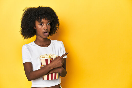 Teen girl with popcorn, cinema concept on yellow backdrop pointing to the side