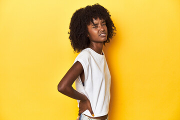 Teen girl in white tank top, yellow studio background suffering a back pain.