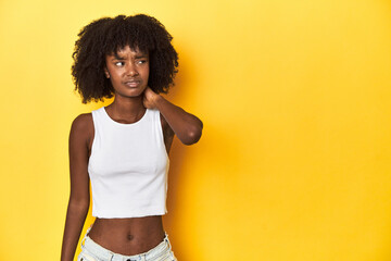 Teen girl in white tank top, yellow studio background touching back of head, thinking and making a choice. - obrazy, fototapety, plakaty