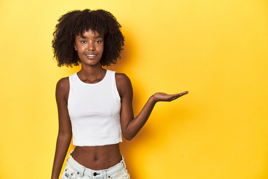 Teen girl in white tank top, yellow studio background showing a copy space on a palm and holding another hand on waist.