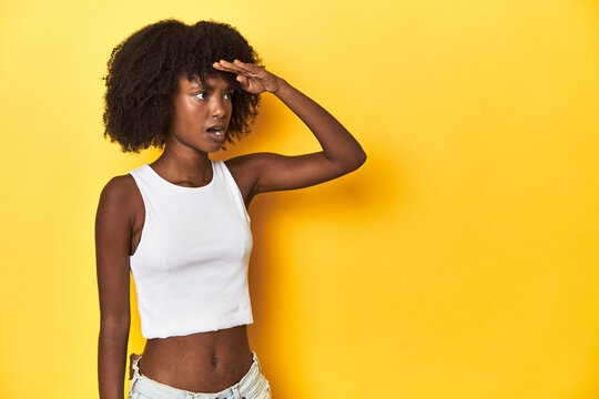 Teen girl in white tank top, yellow studio background looking far away keeping hand on forehead.