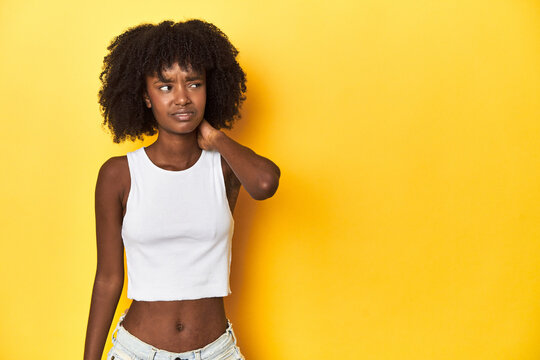 Teen girl in white tank top, yellow studio background touching back of head, thinking and making a choice.
