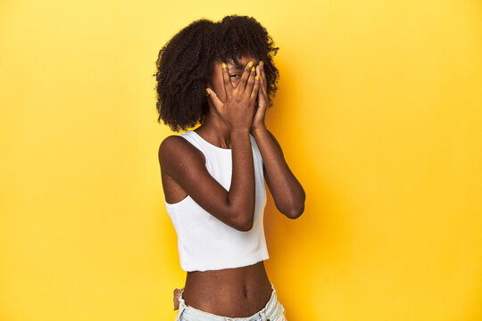 Teen girl in white tank top, yellow studio background blink through fingers frightened and nervous.