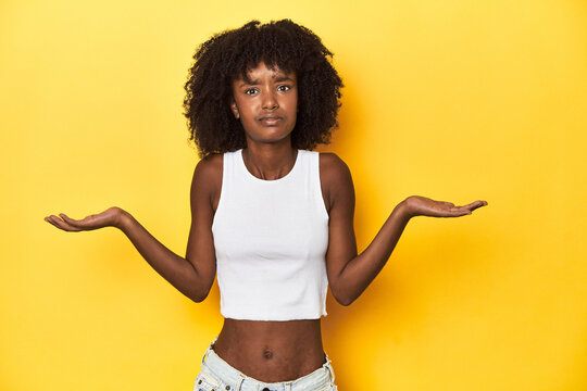 Teen girl in white tank top, yellow studio background doubting and shrugging shoulders in questioning gesture.