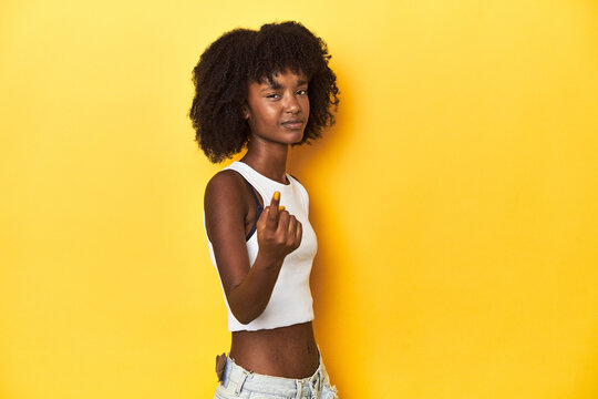 Teen girl in white tank top, yellow studio background pointing with finger at you as if inviting come closer.