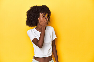 Teen girl in classic white T-shirt, yellow studio backdrop blink at the camera through fingers,...