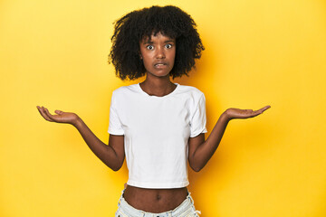 Teen girl in classic white T-shirt, yellow studio backdrop confused and doubtful shrugging...