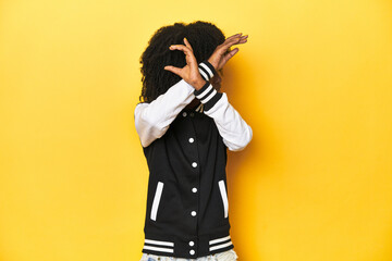 Teen girl in baseball jacket, yellow studio background keeping two arms crossed, denial concept.