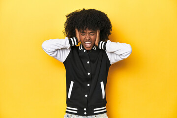 Teen girl in baseball jacket, yellow studio background covering ears with hands trying not to hear...