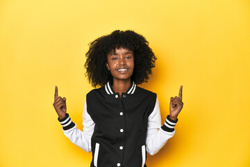 Teen girl in baseball jacket, yellow studio background indicates with both fore fingers up showing...