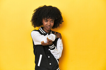 Teen girl in baseball jacket, yellow studio background massaging elbow, suffering after a bad...