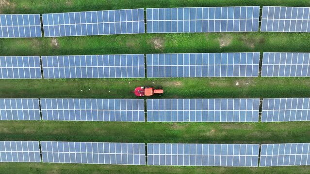 Top down drone view of solar panels in rows with tractor mowing grass between in tropical country. Aerial shot zooming out.