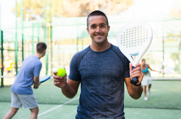 Smiling sporty man standing on outdoor paddle tennis court with racket and ball in hands, ready for...