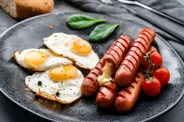 Delicious breakfast with fried eggs, grilled sausages with cheese, tomatoes, greens and spices on plate on dark background. Meat food, selective focus