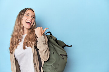 Blonde traveler with a travel backpack points with thumb finger away, laughing and carefree.