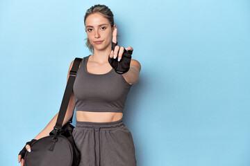 Blonde athlete with gym backpack showing number one with finger.