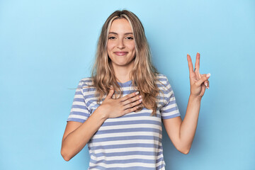 Blonde young caucasian woman in blue studio taking an oath, putting hand on chest.