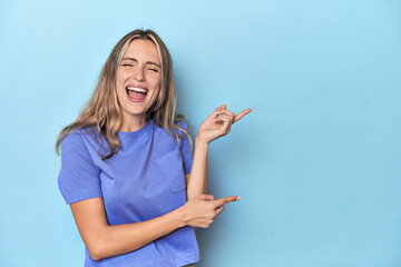 Blonde young caucasian woman in blue studio pointing with forefingers to a copy space, expressing...
