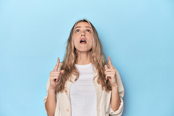Blonde young caucasian woman in blue studio pointing upside with opened mouth.