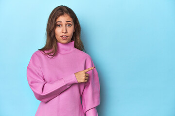 Teen girl in cozy pink sweatshirt on blue pointing to the side