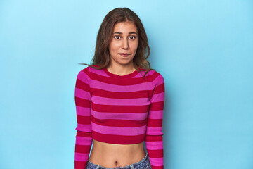 Teen girl in striped pink and red tee on blue shrugs shoulders and open eyes confused. - obrazy, fototapety, plakaty