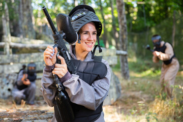 Fototapeta premium Waist-up portrait of happy latin woman in protective paintball equipment standing on outdoor battleground and smiling.