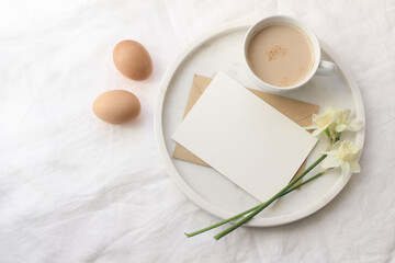 Breakfast spring composition. Mug of latte coffee, bouquet of narcissus, daffodil flowers on marble tray. Blank greeting card mockup. White linen table cloth background. Easter eggs. Flat lay, top