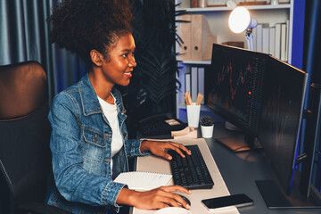 Young African woman trader smiling on happy face, looking on screen with valued stock market chart...