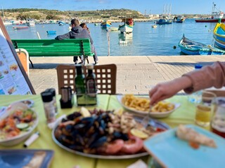 Unidentified people sitting on a bench in front of a table with food in Marsaxlokk, Malta.