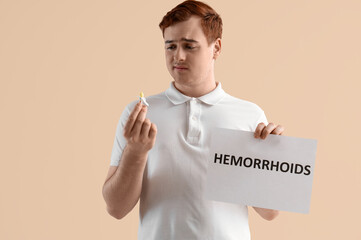 Young man holding paper with word HEMORRHOIDS and rectal suppository on beige background