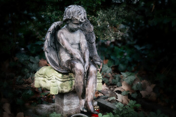 a little angel with wings sits sadly slumped on a stone pillow at a gravestone in a cemetery
