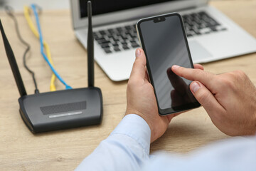 Man with smartphone and laptop connecting to internet via Wi-Fi router at wooden table, closeup