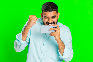 Worried Indian bearded man enthusiastically playing racing drive simulator or shooter video games on smartphone. Happy Arabian guy player celebrate victory isolated on green chroma key background