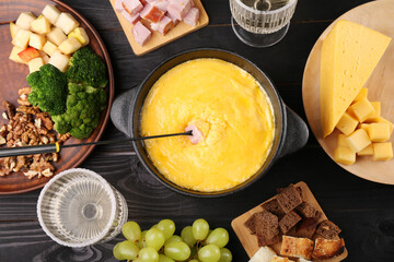 Fondue pot with melted cheese, fork and different products on black wooden table, flat lay