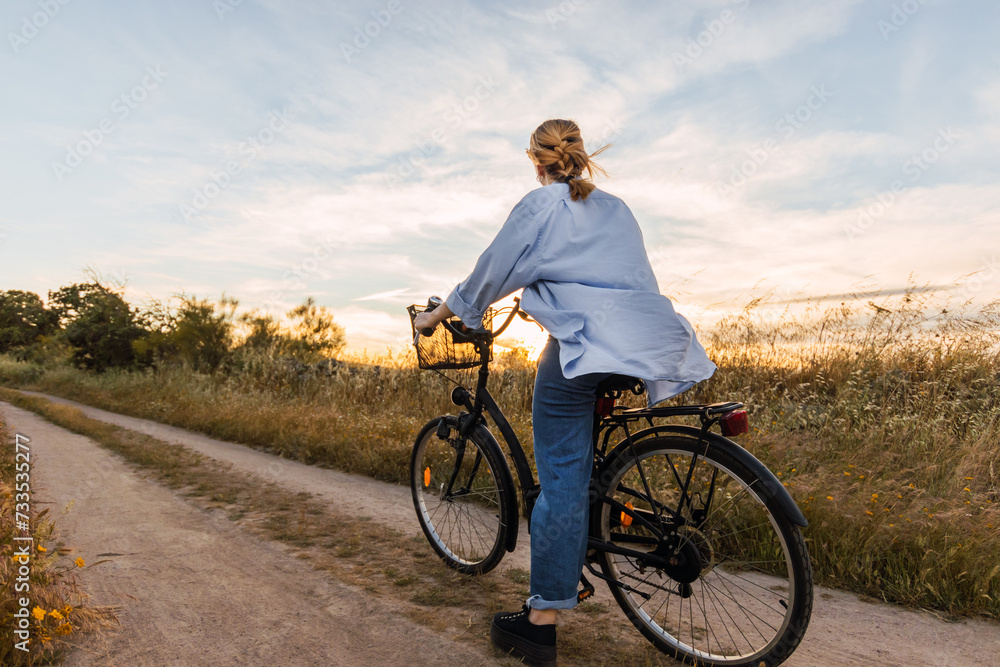 Wall mural back view of a blonde woman on a bike in a field during sunset