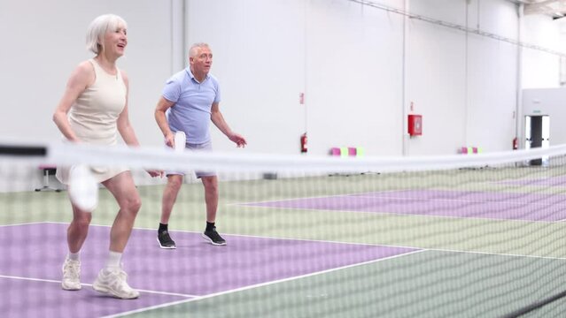 Focused positive aged couple, man and woman, playing friendly doubles pickleball match on indoors court. Healthy active lifestyle of elderly