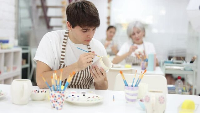 Focused young guy attending pottery class, enthusiastically painting on ceramic cup, creating unique custom design. Creative hobby concept