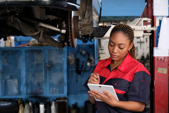 Expert young Black female automotive mechanic worker checks car's wheel brake disc and repairs inspect checklist by tablet at fix garage. Vehicle maintenance service works industry occupation jobs.