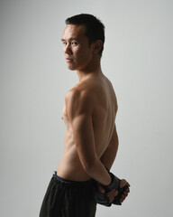 Fototapeta premium Close up portrait of fit asian male model, shirtless with muscles. Wearing gym shorthand boxing gloves, gestural punching pose. Isolated on a white studio background.
