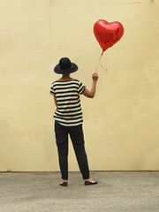 rear view of woman standing alone holding a red heart balloon against wall