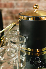Glass drinkware and a black ice bucket with gold accents on a table against a brick background
